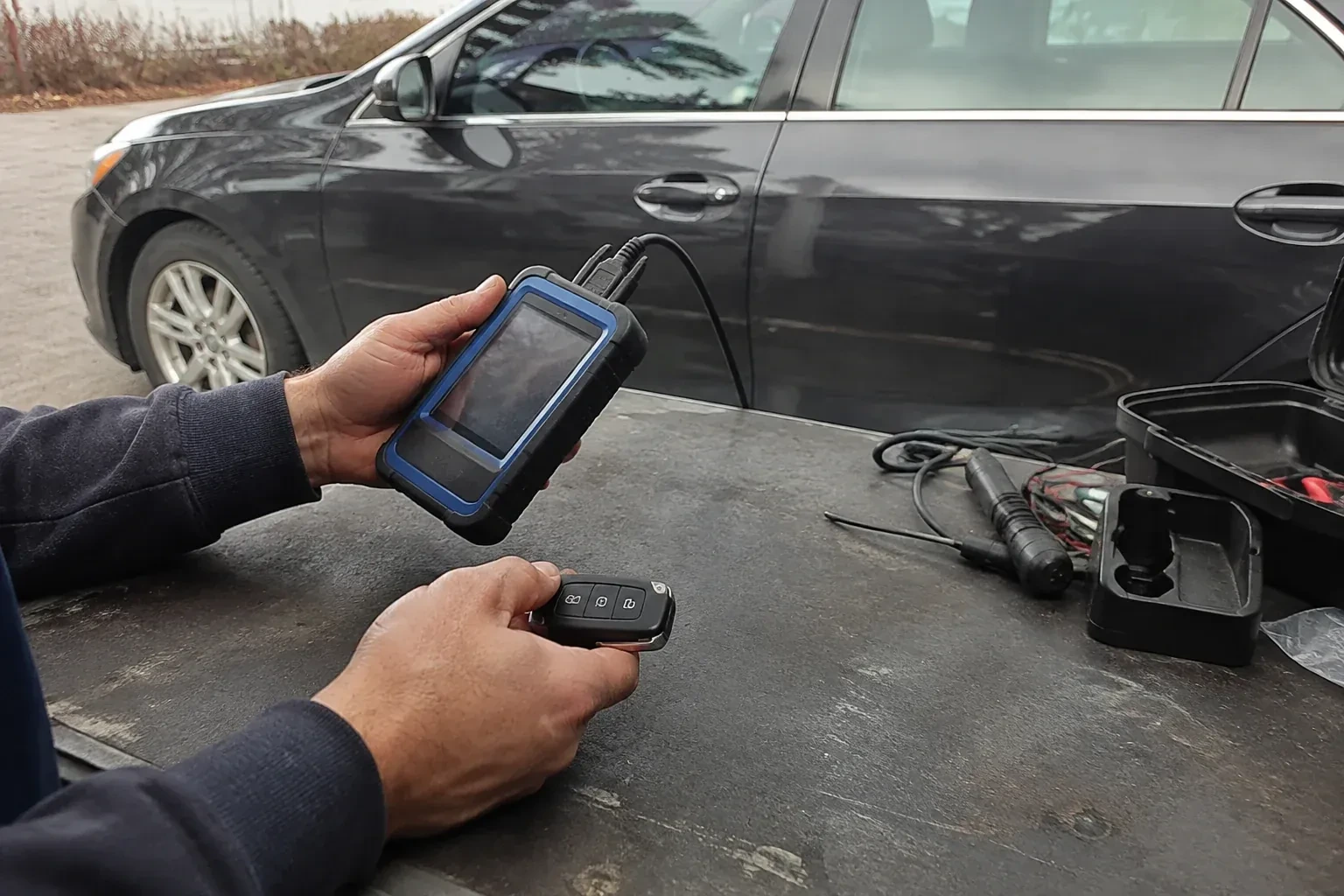 Hands holding a blue-edged electronic device and car-key fob at a worktable beside a parked car. — Transponder Key Programming, Burlington